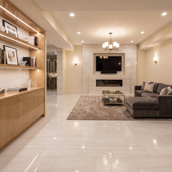 A modern living room with a dark gray sectional sofa, a glass coffee table, a built-in fireplace, and a large light-colored tile floor. A wooden bookshelf with built-in lighting is visible on the left.