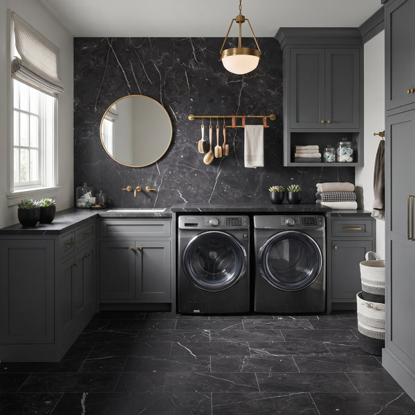A modern laundry room featuring a pair of dark gray front-loading washing machines and a matching sink cabinet against a black marble backsplash. The room is styled with dark gray cabinets, brass hardware, a round gold-framed mirror, and a hanging brass soap dish with utensils.