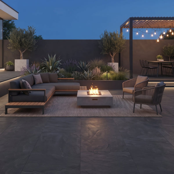 Modern outdoor patio furniture set on a dark stone patio at dusk, featuring a sectional sofa, two armchairs, a rectangular fire pit table, a pergola with string lights, and a built-in grill area.
